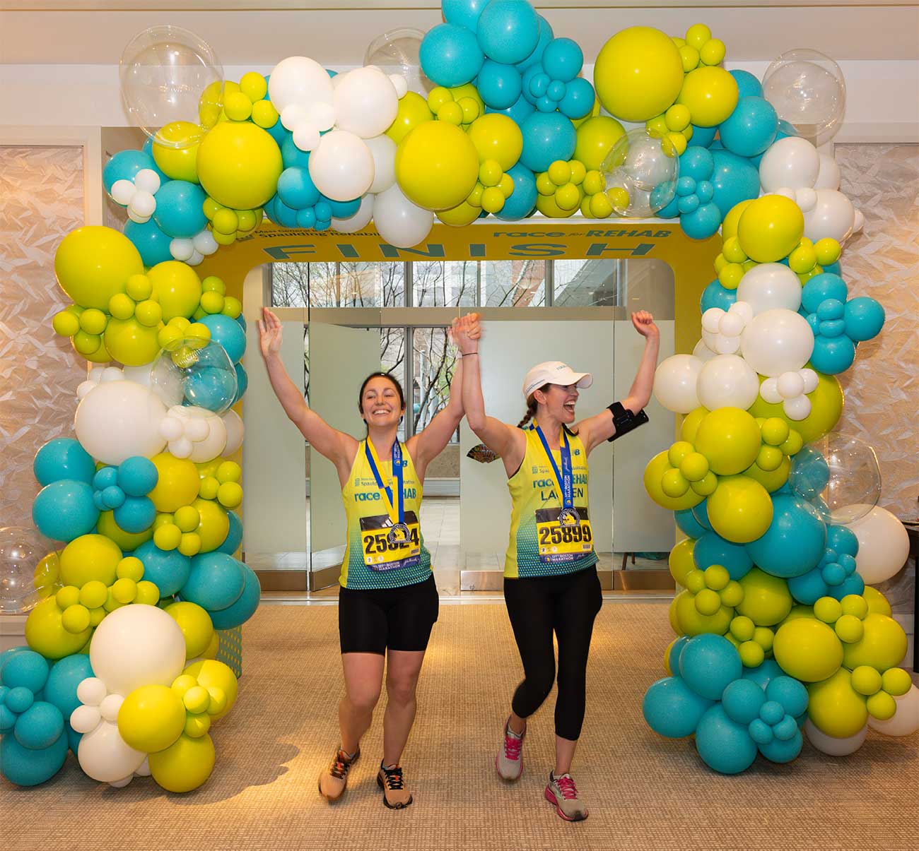 Two women in Race for Rehab shirts grin as they walk, arms raised, under an arch of balloons.