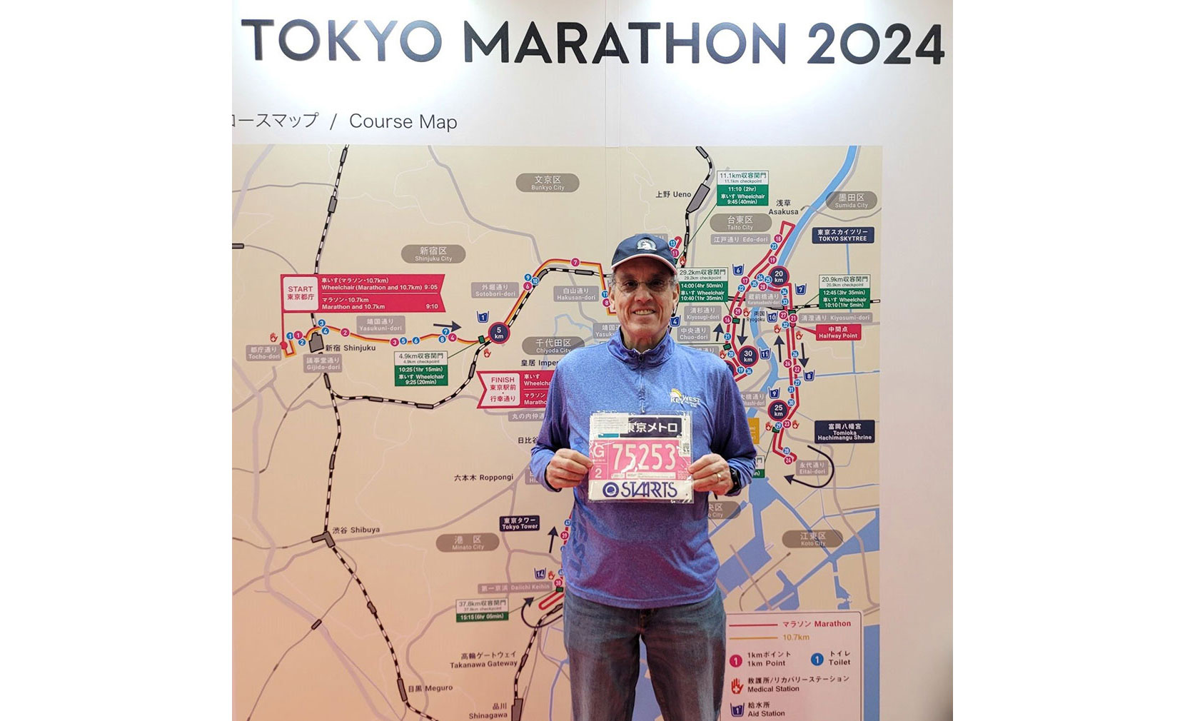 A smiling older man holding up his marathon tag in front of a map of the Tokyo Marathon 2024 course.