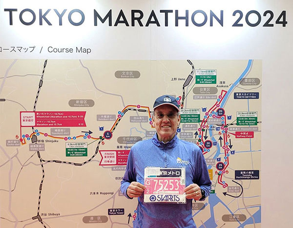 A smiling older man holds up his marathon tag in front of a map of the Tokyo Marathon 2024 course.