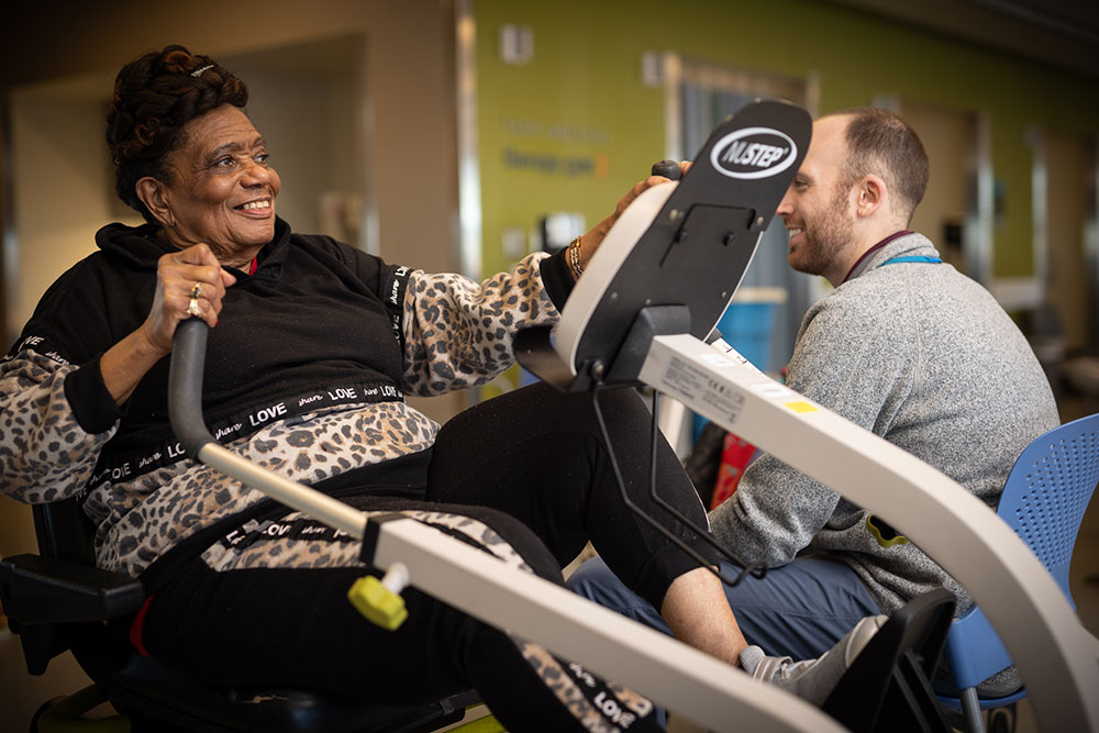 An older woman uses a rowing machine, assisted by her physical therapist.