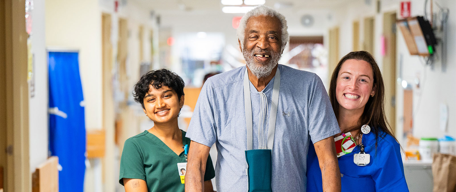 An elderly patient smiles as he walks down the hospital hall, assisted by two physical therapists.