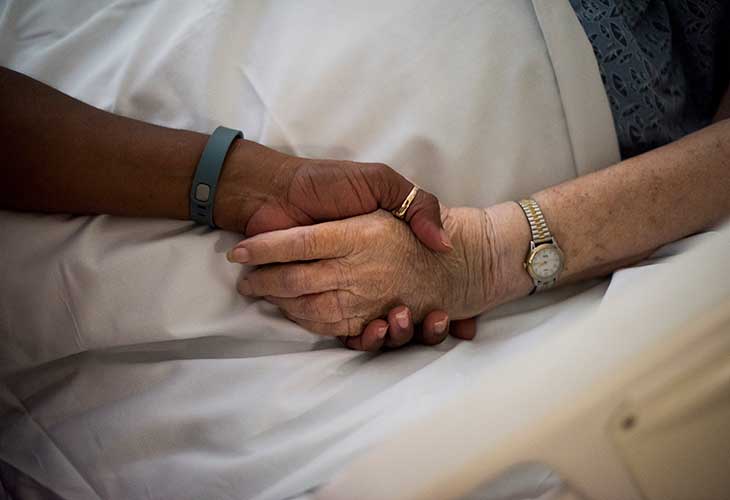 Two clasped hands, an older woman's and a younger woman's, rest on the sheets of a hospital bed.