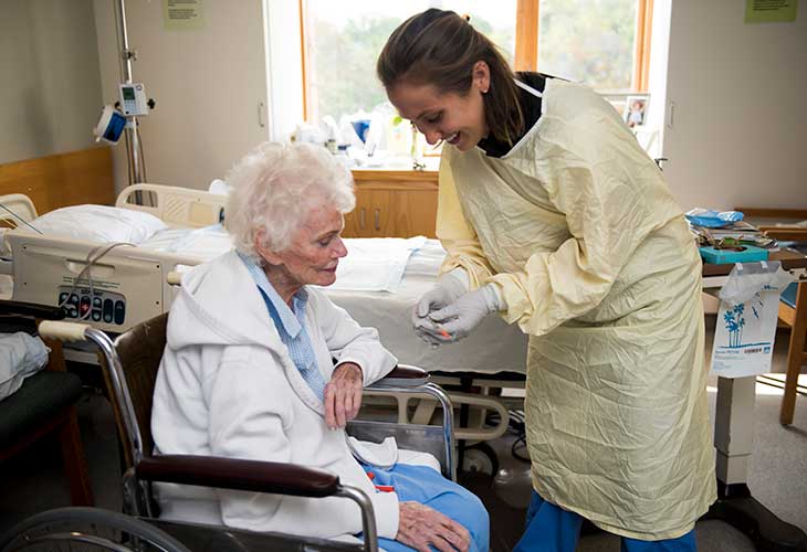 A smiling nurse shows a thermometer reading to an older woman in a wheelchair.