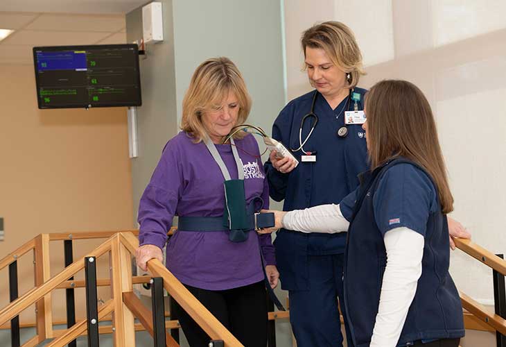 Two nurses in scrubs help a patient, who's wearing a chest monitor and walking down the stairs.