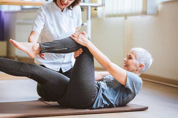 An older woman does an exercise on a mat with the help of a physical therapist.