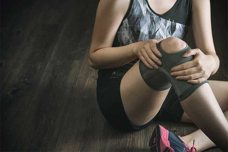A young woman in athletic gear and a knee brace sits on the floor, holding her knee.
