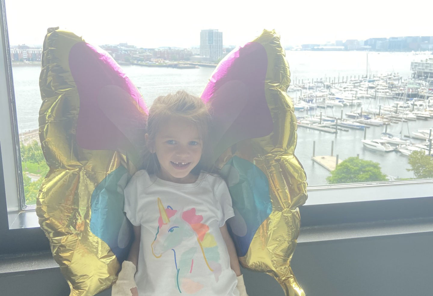 Addy, a small girl wearing rainbow wings, stands at a window overlooking Boston Harbor.