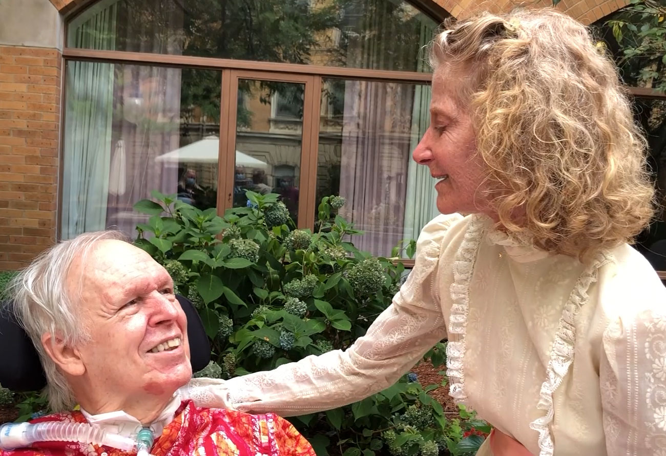 Randy, an older man in a wheelchair, and Catherine, an older woman, share a moment on their patio.