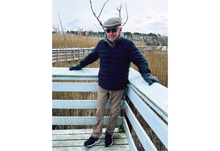 George, an older white man in a coat and hat, leans against the railing of a deck.
