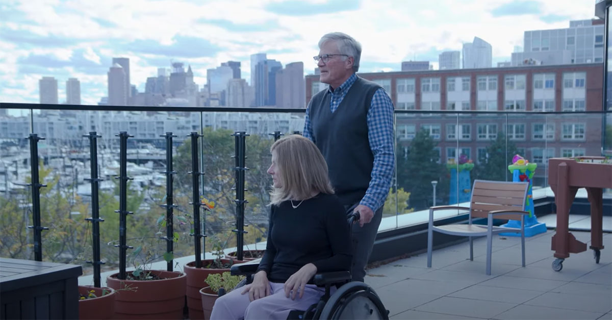 Michele (in a wheelchair) and her husband on a balcony overlooking Boston Harbor.