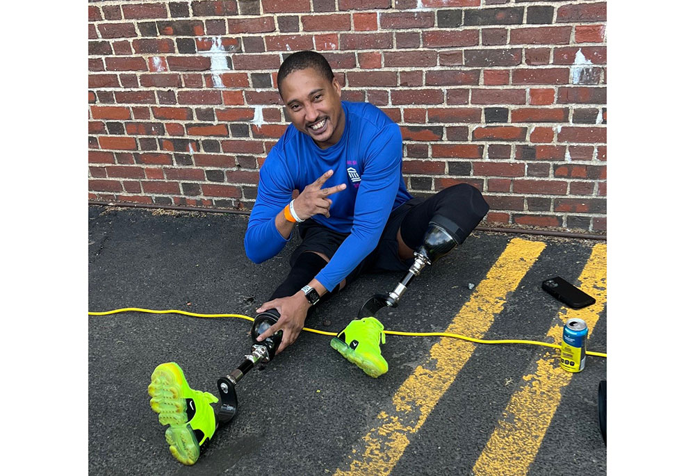 A smiling man stretching while wearing two lower-leg prostheses with neon yellow sneakers.