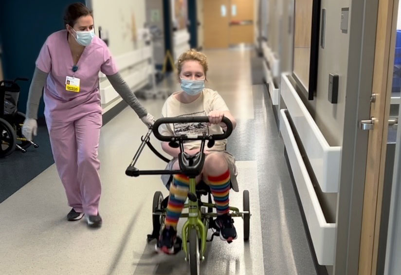 A 12-year-old girl in rainbow socks cycles an adaptive tricycle down a hospital hallway.