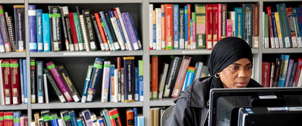 A woman reads intently from a computer screen with a wall of library shelves behind her.